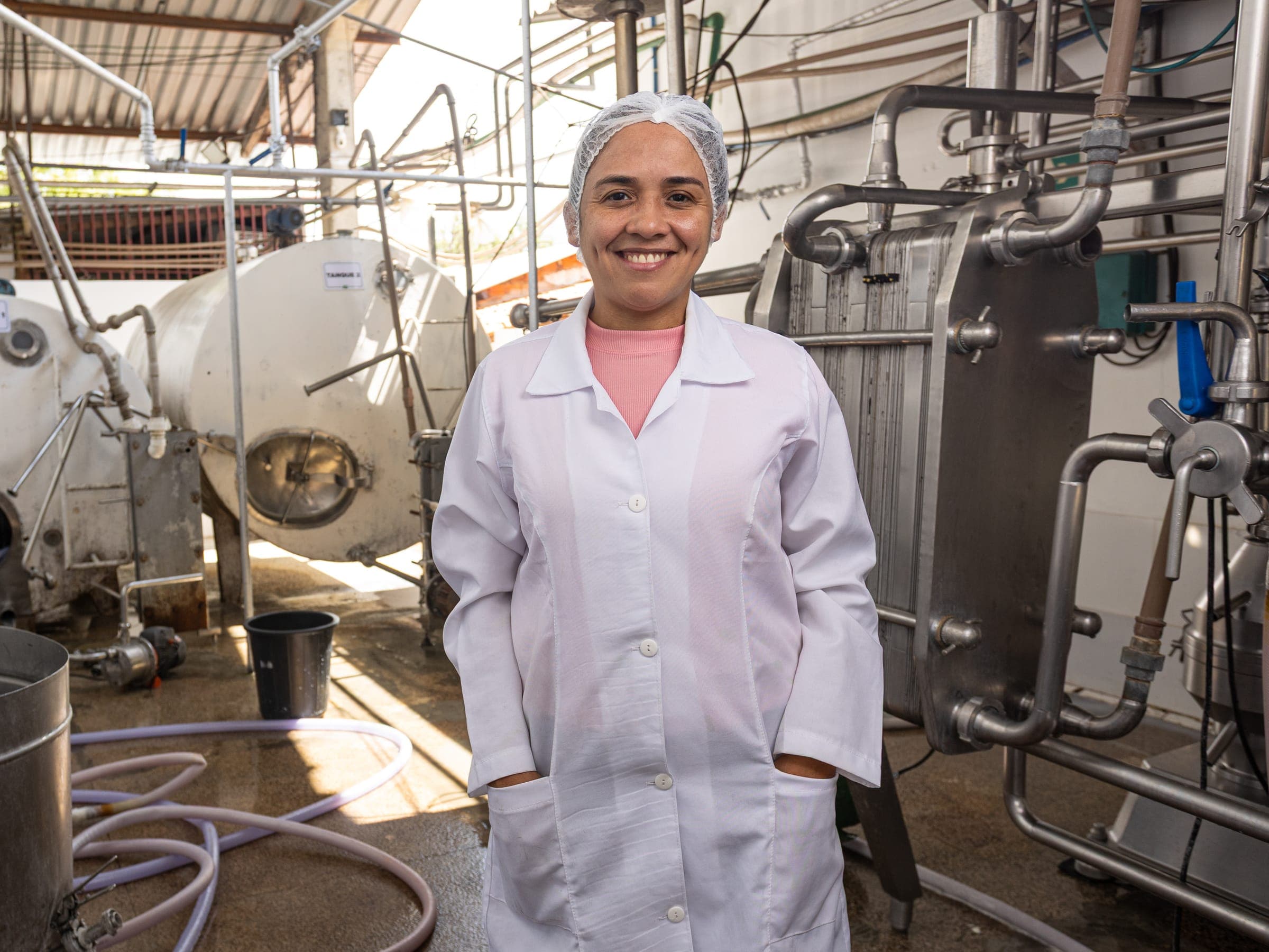 Funcionária de jaleco sorrindo na área de recepção do leite, entre tanques de inox